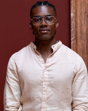 A young man with short, braided hair and glasses is wearing a light-colored shirt and standing in front of a red wall.