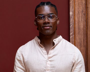 A young man with short, braided hair and glasses is wearing a light-colored shirt and standing in front of a red wall.