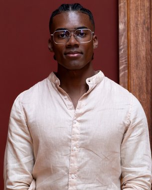A young man with short, braided hair and glasses is wearing a light-colored shirt and standing in front of a red wall.
