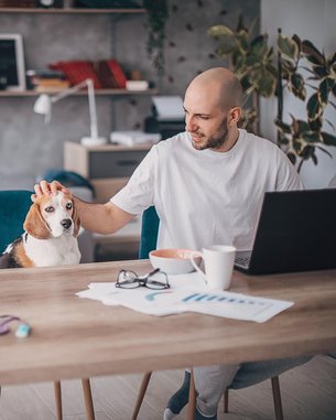 A bald man is petting a Beagle while working at a table with a laptop and a cup of coffee. There are also documents and a pair of glasses on the table.