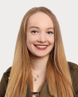 A young woman with long blonde hair and a friendly smile is wearing a green jacket. The background is neutral and bright.