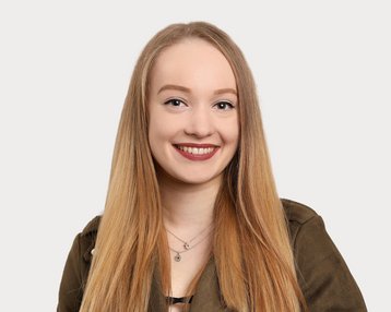 A young woman with long blonde hair and a friendly smile is wearing a green jacket. The background is neutral and bright.