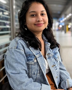 A young woman with long, dark hair is sitting on a bench, wearing a denim jacket and smiling in a modern interior.