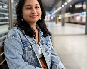 A young woman with long, dark hair is sitting on a bench, wearing a denim jacket and smiling in a modern interior.