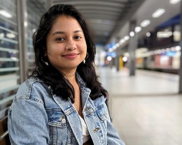 A young woman with long, dark hair is sitting on a bench, wearing a denim jacket and smiling in a modern interior.