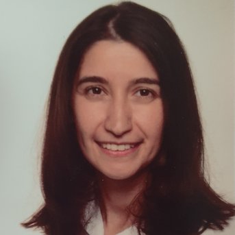 A woman with long, dark hair and a friendly smile is wearing a light-colored shirt. The background is neutral and bright.