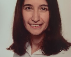 A woman with long, dark hair and a friendly smile is wearing a light-colored shirt. The background is neutral and bright.