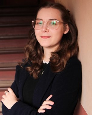 A young woman with curly hair and glasses leans against a wall, looking at the camera. She is wearing a black sweater and a dark blue coat. In the background, stair steps are visible.
