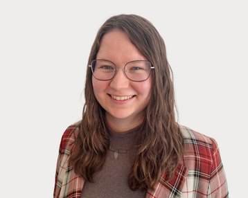 A smiling person with long, curly hair is wearing a checkered jacket and glasses. The background is neutral.