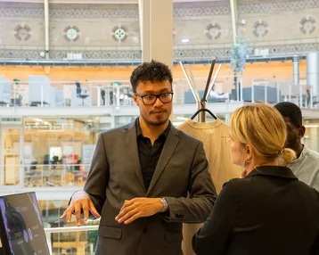 A man in a black suit is speaking with a woman in a modern office. In the background, workstations and plants are visible.