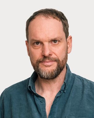 A man with a beard and short hair is wearing a blue shirt. He is looking directly at the camera against a neutral background.