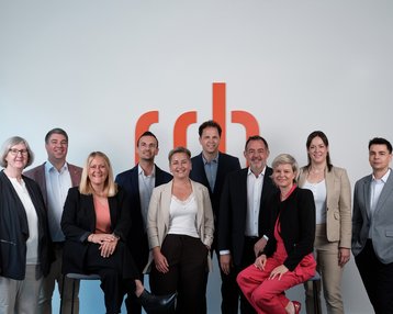 A group of ten people stands in front of a wall with the "rdb" logo. They are dressed in formal attire and smiling at the camera.