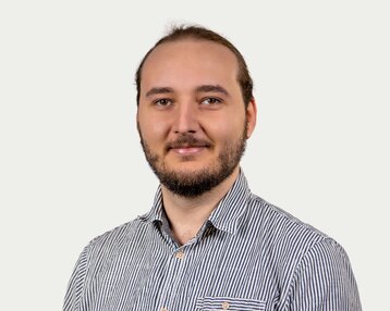 A man with a beard and long hair is wearing a striped shirt and smiling at the camera against a neutral background.