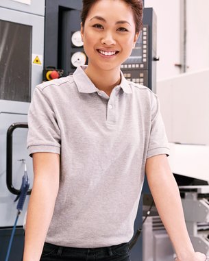 A smiling woman stands at a table in a workshop, surrounded by tools and machines. She is wearing a gray polo shirt and has her arms resting on the table.