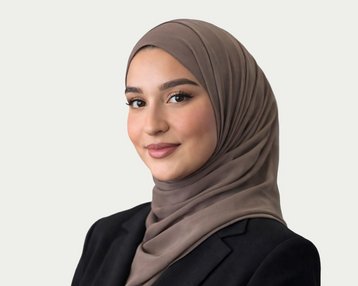 A young woman wearing a brown hijab and a black blazer, smiling at the camera. The background is kept neutral.