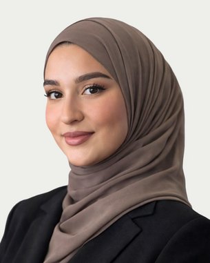A young woman wearing a brown hijab and a black blazer, smiling at the camera. The background is kept neutral.