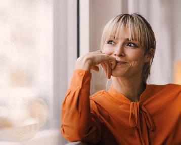 A woman with blonde hair and an orange top sits thoughtfully by the window, resting her chin on her hand.