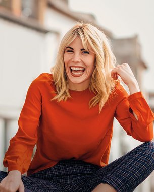 A smiling woman with blonde hair is wearing a red top and sitting with her legs crossed. Blurry buildings can be seen in the background.