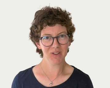 Woman with curly brown hair and glasses, wearing a black top. She is looking directly at the camera.