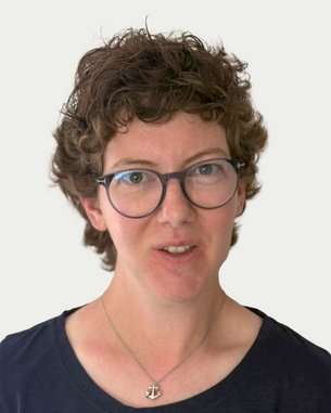 Woman with curly brown hair and glasses, wearing a black top. She is looking directly at the camera.