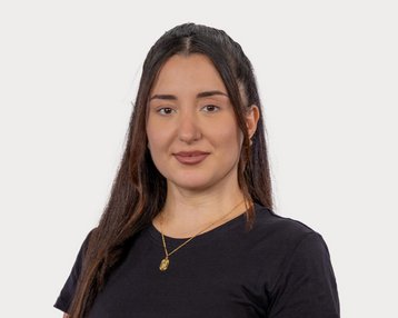 A young woman with long, dark hair is wearing a black T-shirt and a gold necklace. She is looking directly at the camera.