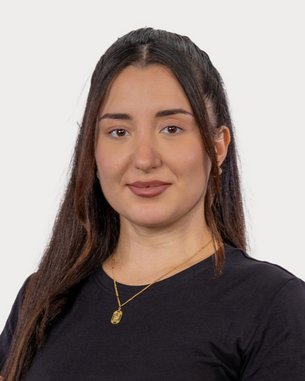 A young woman with long, dark hair is wearing a black T-shirt and a gold necklace. She is looking directly at the camera.