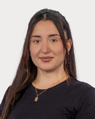 A young woman with long, dark hair is wearing a black T-shirt and a gold necklace. She is looking directly at the camera.