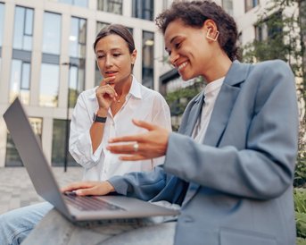 Two women are sitting outside, looking at a laptop. One woman is pointing at the screen while the other appears to be deep in thought. Both are dressed in modern clothing and are in an urban setting.