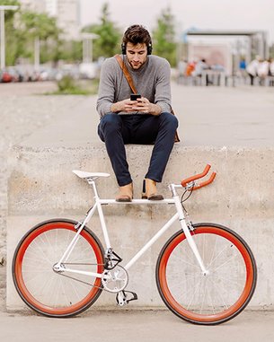 A man is sitting on a concrete wall, looking at his smartphone. Next to him is a bicycle with a white frame and orange tires.
