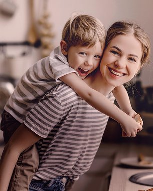 A smiling woman is carrying a boy on her back. Both are wearing striped T-shirts. In the background, kitchen appliances and utensils are visible.