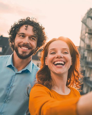 A man with curly hair and a woman with long, red hair are smiling and taking a selfie against an urban backdrop. Both are dressed casually and appear relaxed.