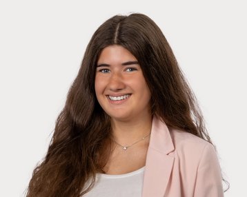 Young woman with long, curly hair, smiling in a light blazer and a white top. Neutral background.