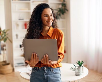 A woman with curly hair is holding a laptop and smiling. She is wearing an orange shirt and standing in a bright room filled with plants and a table.