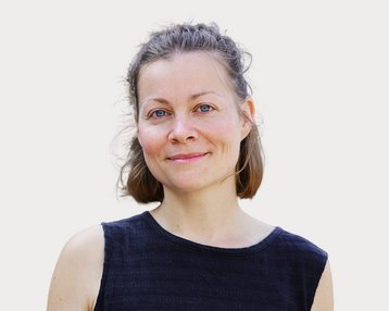 A woman with short brown hair and blue eyes is wearing a black top and smiling warmly at the camera.