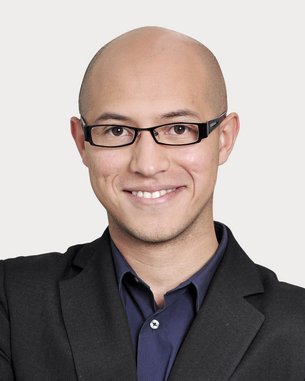 Man with a bald head and glasses, wearing a black suit and a dark shirt, smiles warmly and stands with his arms crossed.