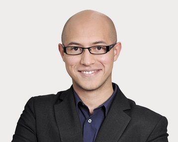 Man with a bald head and glasses, wearing a black suit and a dark shirt, smiles warmly and stands with his arms crossed.