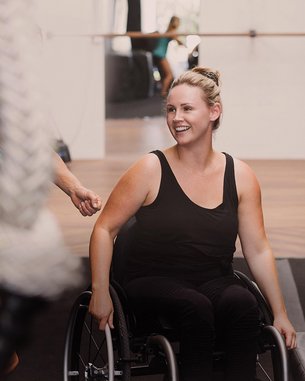 A woman in a wheelchair smiles as she sits in a training room. In the background, other people and exercise equipment can be seen.