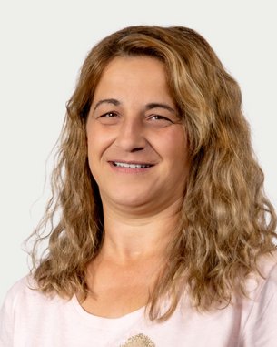 A woman with curly brown hair smiles warmly at the camera. She is wearing a light-colored T-shirt and has a neutral background.