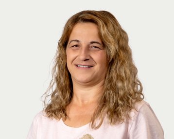 A woman with curly brown hair smiles warmly at the camera. She is wearing a light-colored T-shirt and has a neutral background.