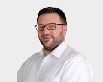 A man with glasses and a beard is wearing a white shirt. He is smiling and standing slightly sideways in front of a neutral background.