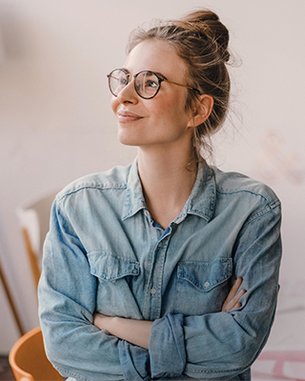 Young woman with glasses and a casual denim shirt, looking thoughtfully with her arms crossed. In the background, furniture and a lamp are visible.